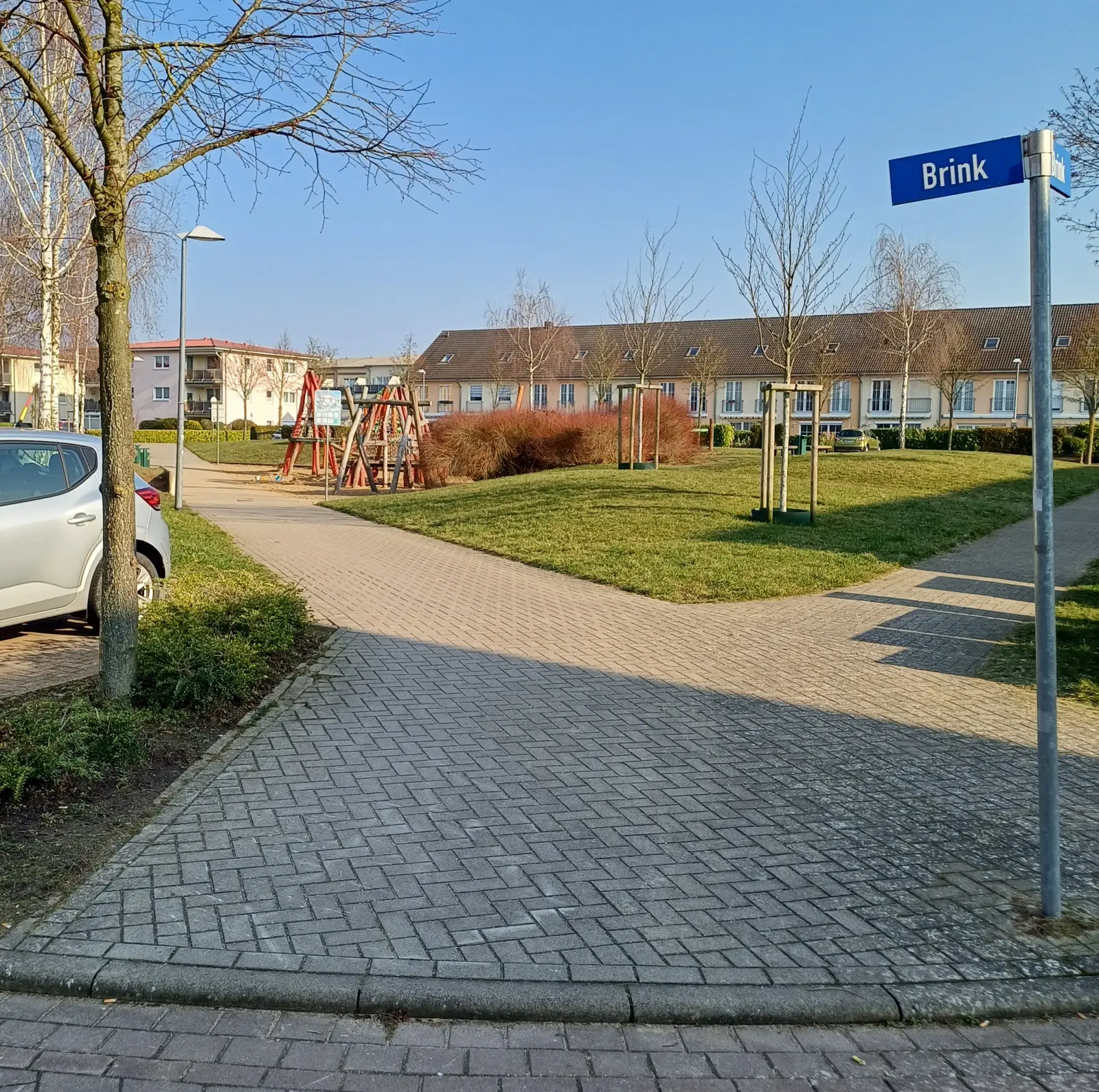 Gepflasterter Weg mit Straßenschild „Brink“, dahinter Rasen und Reihenhäuser